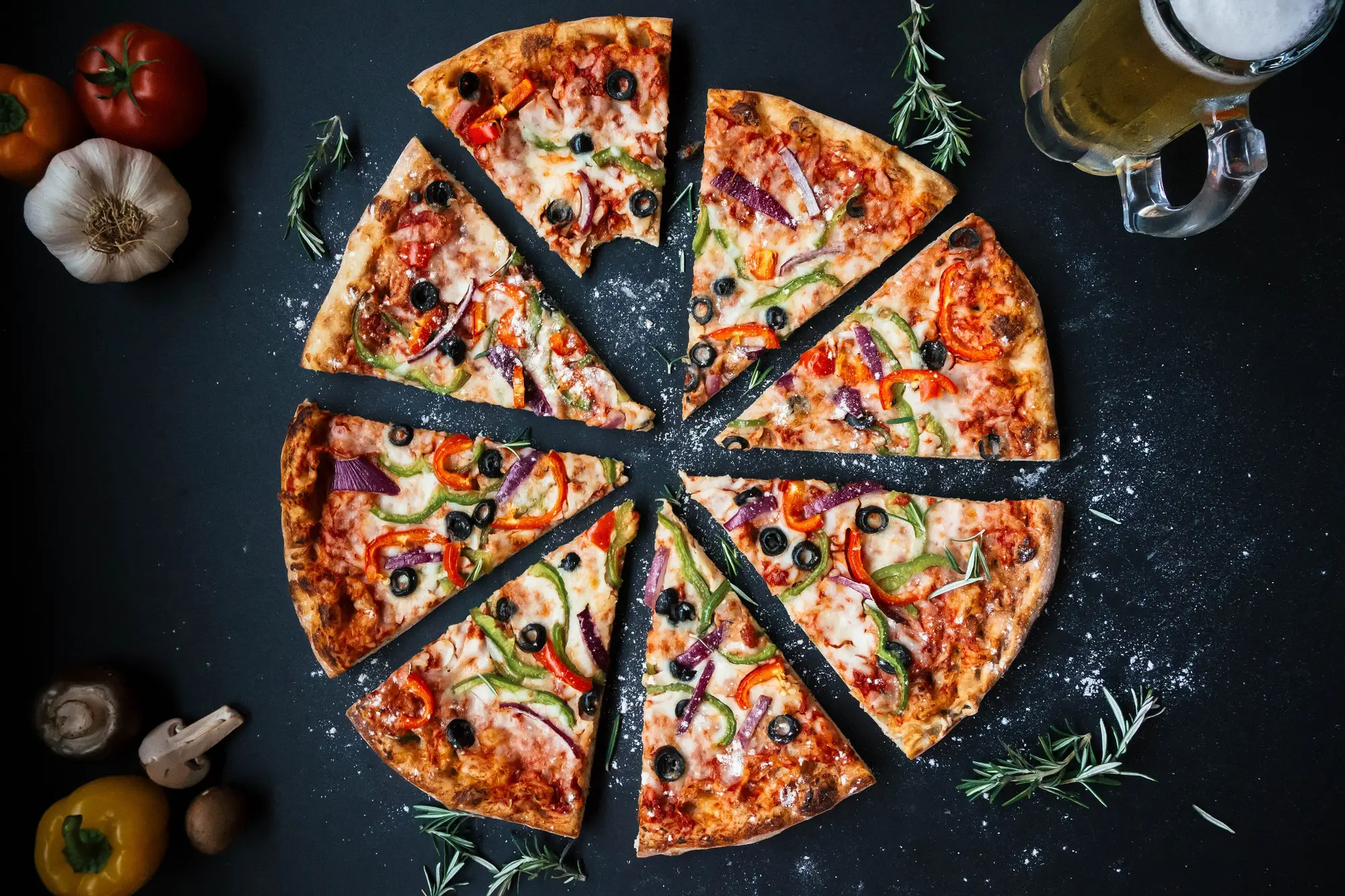 Sliced vegetable pizza on a dark table with garlic, tomatoes and rosemary